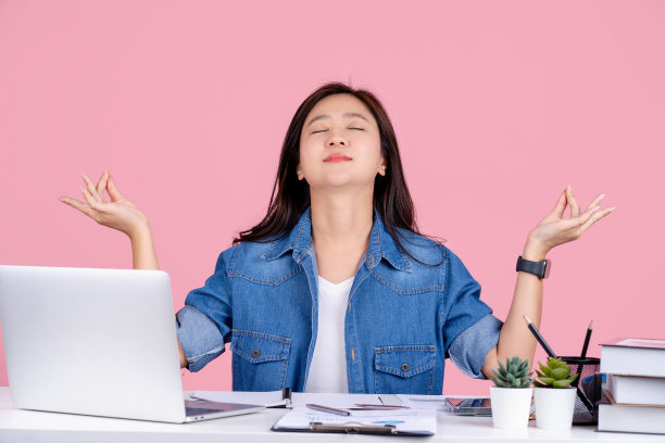 年轻的亚洲商业女性穿着休闲衬衫坐在白色办公桌与pc笔记本电脑隔离在粉彩背景工作室工作。她在做瑜伽，手拉手，放松，冥想，试图平静下来。图片下载