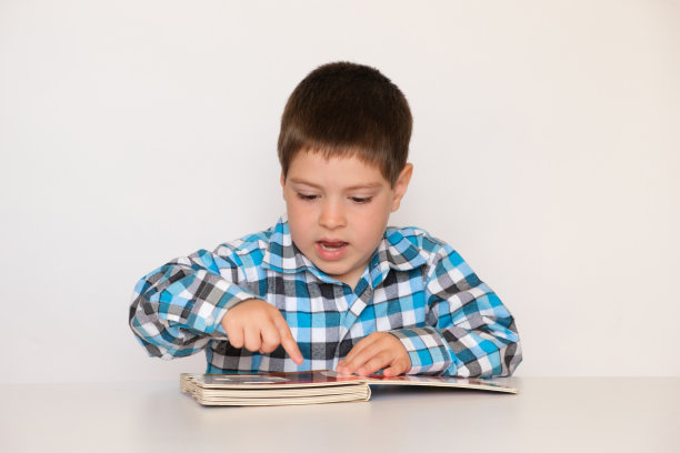 一个4岁的男孩看着一本厚厚的硬纸板，白色背景上有图片的书。图片下载