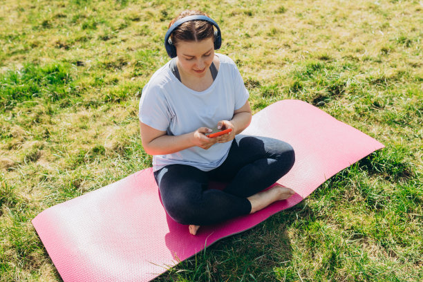 美丽的年轻红发女人放松与智能手机在瑜伽垫上训练后。快乐的女孩与耳机听音乐或浏览互联网。运动或瑜伽的概念图片下载