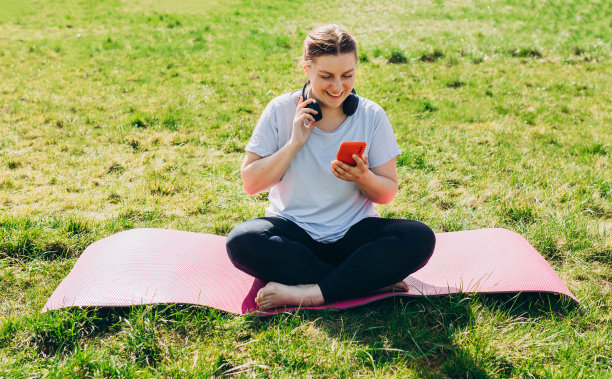 美丽的年轻红发女人放松与智能手机在瑜伽垫上训练后。快乐的女孩与耳机听音乐或浏览互联网。运动或瑜伽的概念图片下载