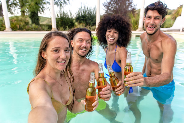 夏季泳池派对!一群快乐的多种族朋友站在水里，喝啤酒，庆祝炎热的假期。人们在欢乐时光自拍。生活方式的概念图片下载