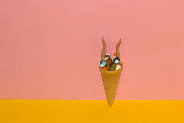 圆锥形冰淇淋上有抽象的彩色分子球，手从球里伸出来，黄粉色背景上的创意夏季设计图片下载