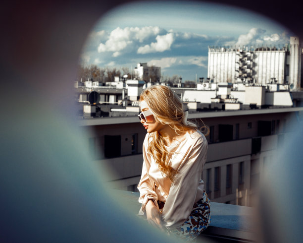 年轻女子的肖像与长金发穿着米色丝绸衬衫，印花长裤，太阳镜，坐在屋顶。图片下载