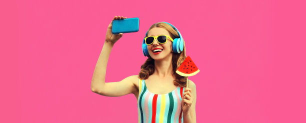 夏天的肖像快乐快乐的笑年轻女子自拍与智能手机在耳机听音乐多汁棒棒糖或冰淇淋形状的西瓜片在粉红色的背景图片下载