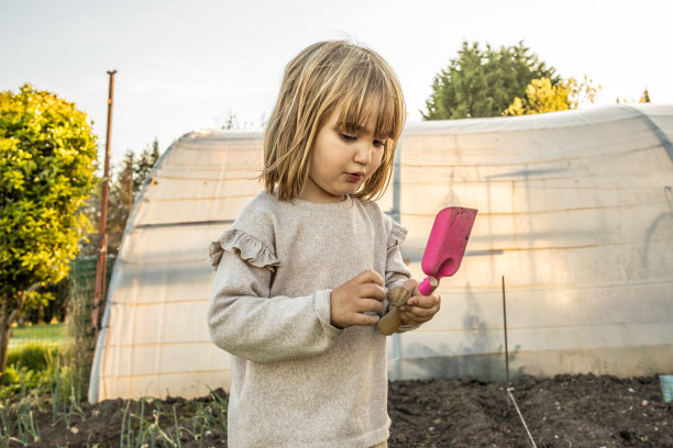 一个女孩拿着铲子挖土，身后是一个温室图片下载
