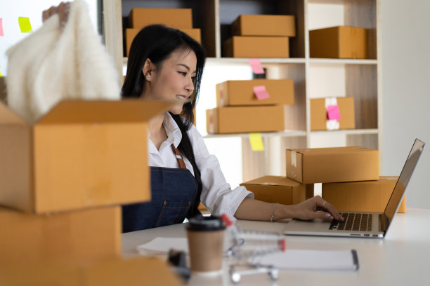 一名女性电子商务小企业主正在使用笔记本电脑检查网上订单，并将它们打包运送。图片下载