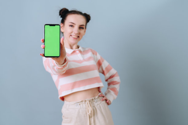 一个梳着发髻的现代女孩在空墙上展示一个绿色屏幕的手机。专注于手机图片下载