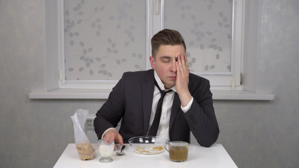 一个穿着西装的昏昏欲睡的年轻人吃麦片，喝咖啡。年轻的商人在家里的厨房里。图片下载