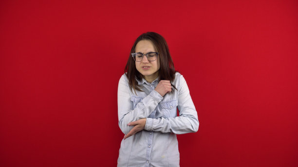 一个戴眼镜的年轻女子手肘酸痛，她把手肘握在手里。在红色背景上拍摄。图片下载