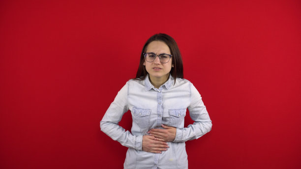 一位戴眼镜的年轻女士胃痛，她把眼镜拿在手里。在红色背景上拍摄。图片下载