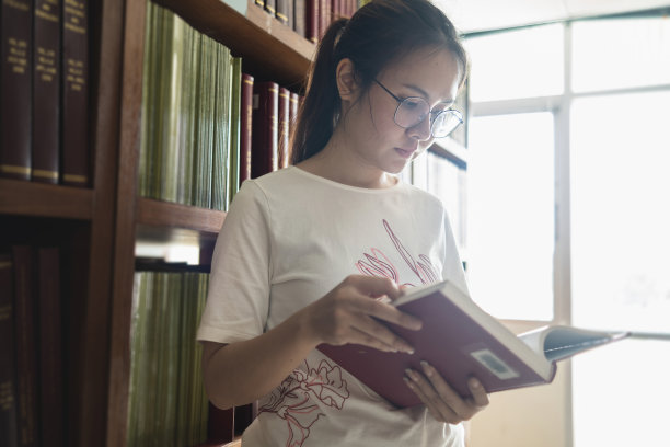 一位亚洲妇女站在图书馆的书架走廊上看书。图片下载