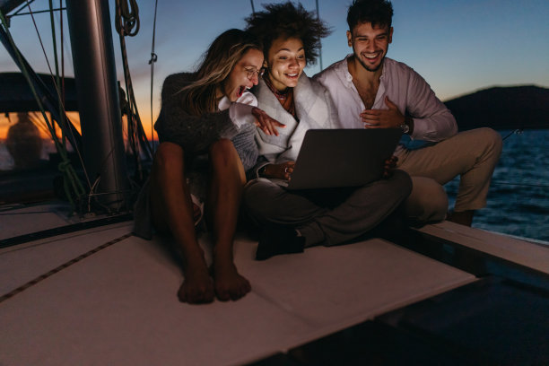 三个朋友晚上坐在帆船的甲板上，用笔记本电脑在流媒体平台上看电影，面带微笑图片下载