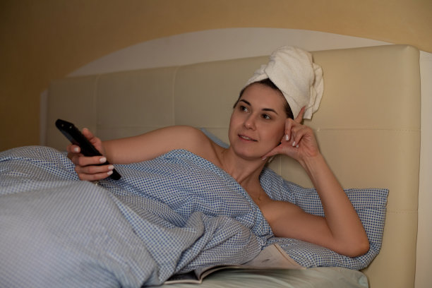 头发上裹着毛巾，躺在床上，看杂志，看电视，在酒店房间里用遥控器图片下载