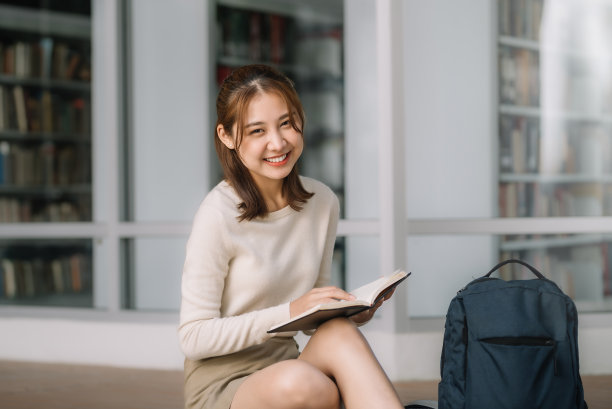年轻的亚洲女学生坐着，拿着书看着相机。图书馆背景图片下载