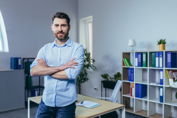 年轻成功商人的肖像，男人在明亮的办公室看着相机，微笑着双臂交叉图片下载
