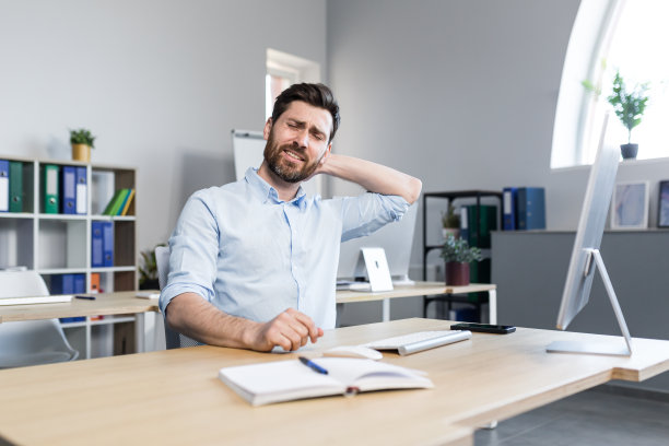 一个疲惫的年轻人在电脑前工作，坐在办公室的桌子前，感到脖子疼痛，按摩他的脖子，放松。图片下载