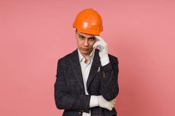 年轻的男工程师戴着安全帽，穿着夹克，在粉红色的背景下思考图片下载