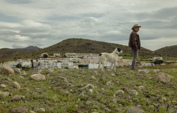 戴着牛仔帽的成年男子和他的狗站在田野上，城市在天空的背景下图片下载