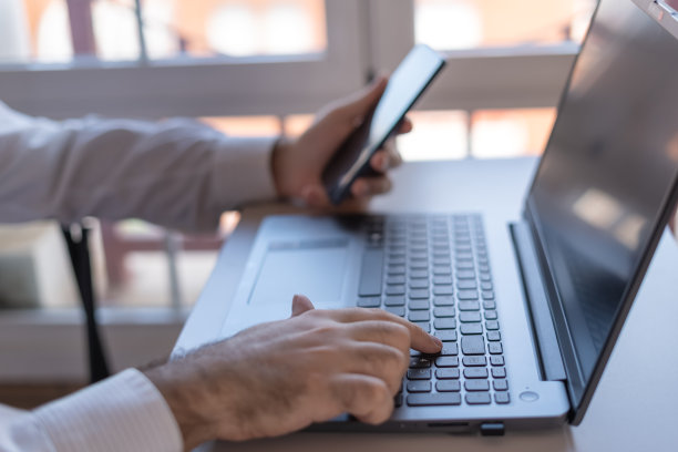 男人在远程办公的时候，一边在笔记本电脑上打字，一边查看手机。图片下载