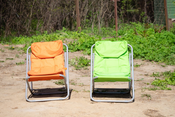 橙色和绿色的休闲折叠式旅游椅，背景是棕色的土地，绿色的草地，阳光下的铁网栅栏图片下载