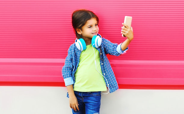 幸福微笑的小女孩的肖像自拍与智能手机和耳机听音乐在城市的粉红色背景图片下载