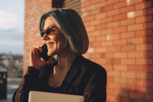 蓝色头发的女人站在外面的砖墙上，微笑着用智能手机聊天。移动电话。移动通信。说话的人。商业的女人。图片下载