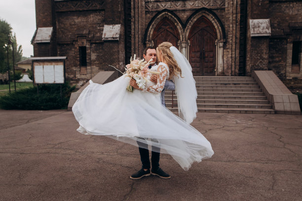 新郎把新娘抱在怀里。美丽的新婚夫妇在古老的基督教教堂附近图片下载