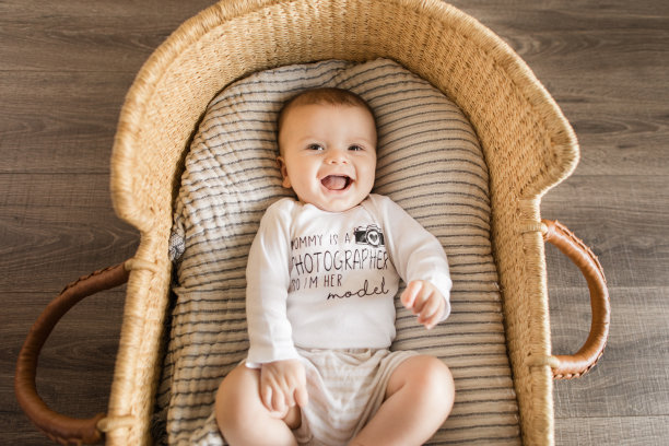 一个19周大的微笑小男孩穿着一件写着“妈妈是摄影师，所以我是她的模特”的白色衣服，躺在一个舒适的奶油色条纹棉毯上，在海草摩西篮子里图片下载