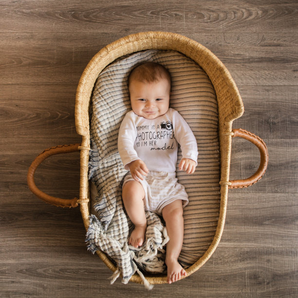 一个19周大的微笑小男孩穿着一件写着“妈妈是摄影师，所以我是她的模特”的白色衣服，躺在一个舒适的奶油色条纹棉毯上，在海草摩西篮子里图片下载