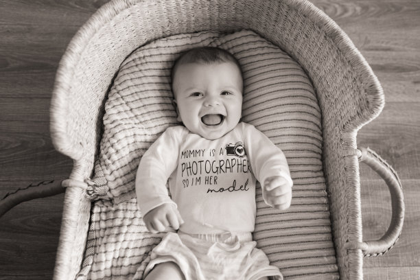 一个19周大的微笑小男孩穿着一件写着“妈妈是摄影师，所以我是她的模特”的白色衣服，躺在一个舒适的奶油色条纹棉毯上，在海草摩西篮子里图片下载