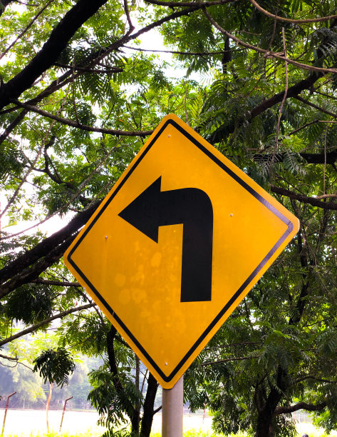 黄色交通标志以绿树为背景左转图片下载