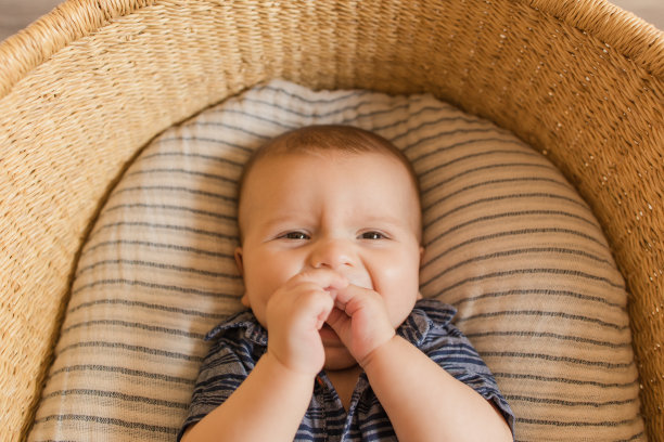 一个21周大的小男孩微笑着，穿着蓝色牛仔装和棕色沙滩凉鞋，躺在一个舒适的奶油条纹棉毯子在海草摩西篮子图片下载