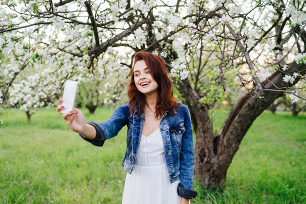年轻漂亮的微笑白人妇女在牛仔裤夹克和白色连衣裙在一个盛开的春天的树自拍。图片下载