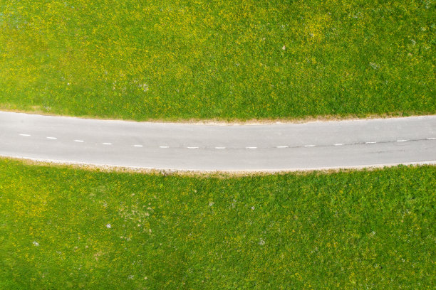 夏日艳阳天绿草地上空旷的道路鸟瞰图。从无人机俯瞰乡村道路、草地和鲜花。美丽的风景与道路在春天。斯洛文尼亚。自然背景图片下载