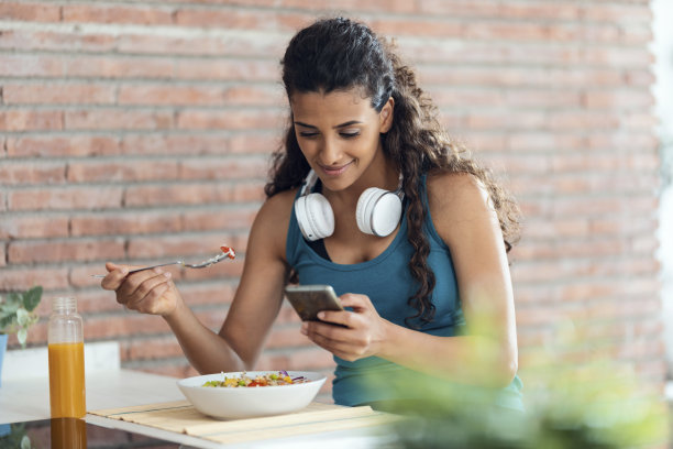 美丽的运动女人吃一碗沙拉，同时在家里的厨房里用手机发短信。图片下载