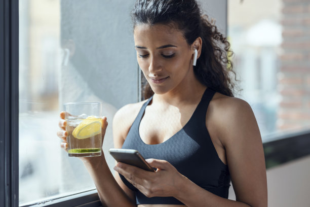 运动女性在家客厅边喝排毒水边使用智能手机。图片下载