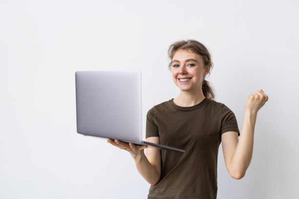 快乐的赢家。年轻开朗的女商人拿着笔记本电脑站在灰色的墙上。看向一边，做出胜利者的手势。图片下载