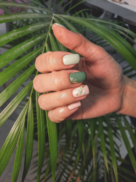 女性的美甲与柔和的颜色和一个分支的设计与叶子的背景上的绿色植物。方钉是绿色、灰色和米色，带有细枝图案和金色亮片。图片下载