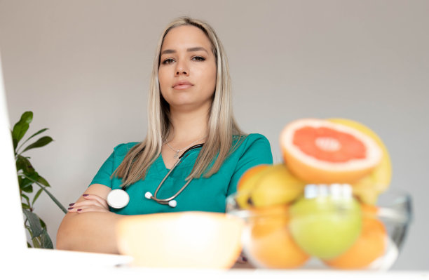 女性营养学家拿着水果在办公桌前工作。微笑的营养师在她的办公室，她正在展示健康的蔬菜和水果，保健和饮食的概念。图片下载