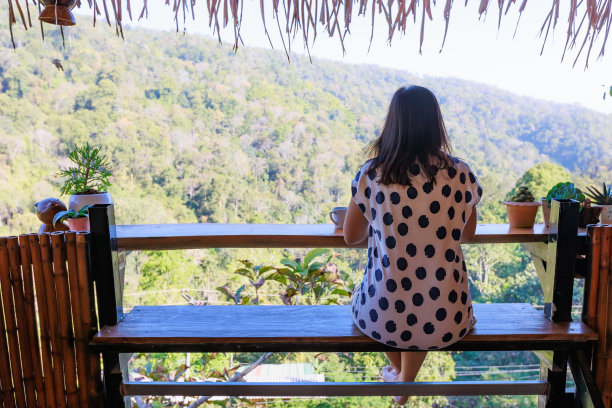 亚洲女人坐在阳台上，在笔记本上写想法，在清新的自然附近与热饮在早上的时间。图片下载