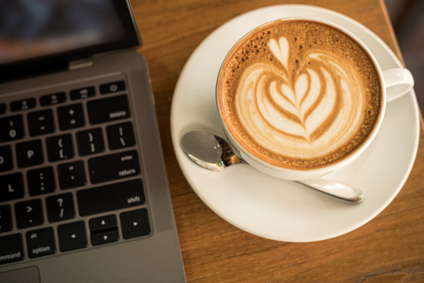 热咖啡拿铁与拿铁艺术奶泡在杯子杯和笔记本电脑在木办公桌办公桌在咖啡馆的咖啡馆，在业务的工作概念图片下载