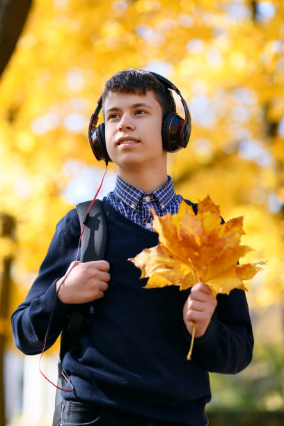 一个十几岁的男孩走在秋天的公园里，戴着耳机听着音乐，阳光明媚的日子里，树上的叶子都泛黄了图片下载