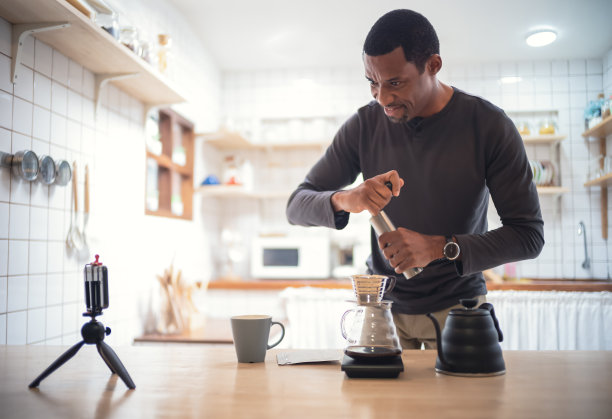 一名非洲裔美国男子在家里的厨房里用智能手机在线观看制作滴滤咖啡的虚拟视频。一名黑人分享专业技能，为他的博客频道滴咖啡。图片下载
