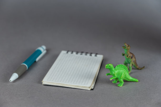 两个恐龙微型模型，笔记本和圆珠笔在灰色的背景。猎物用后腿站立的绿色小图形。弹簧笼笔记本图片下载
