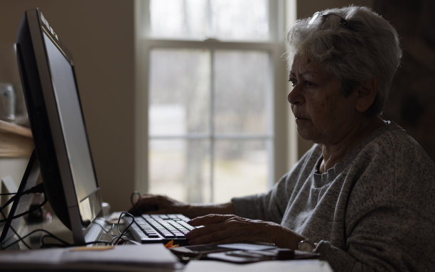 一头银发的老妇人在家庭办公室的个人电脑上工作。图片下载