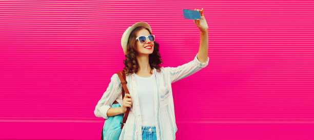 肖像快乐微笑的年轻女子自拍与智能手机戴夏天草帽，背包在粉红色的背景，空白的拷贝空间的广告文本图片下载