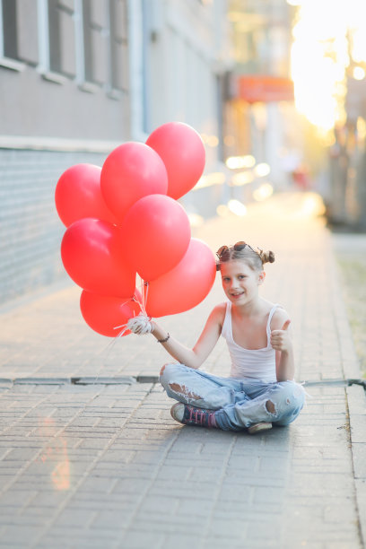 一个穿着牛仔裤和白t恤的女孩坐在人行道上，手里拿着一堆红气球，这是Z一代，孤独的节日图片下载