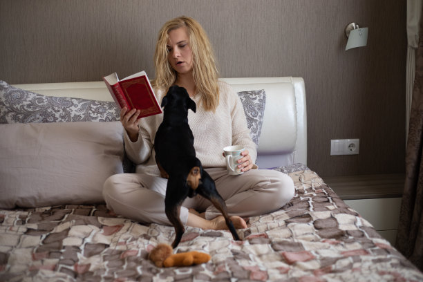 年轻女子的肖像与长金发穿着米色毛衣，长裤，坐在床上，阅读书籍，拿着杯子。图片下载