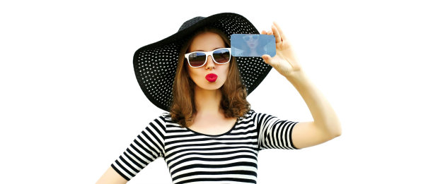 美丽的年轻女子用智能手机自拍吹她的嘴唇与红色的口红发送甜蜜的空气吻戴夏天的草帽，条纹衬衫孤立在白色背景图片下载