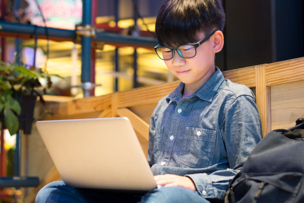 可爱的亚洲男孩盘腿坐在学校，微笑着用笔记本电脑学习网络课程图片下载